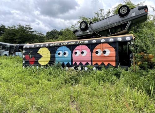 A colorful bus with Pac-Man and ghost characters, surrounded by overgrown grass and a car upside down on top.