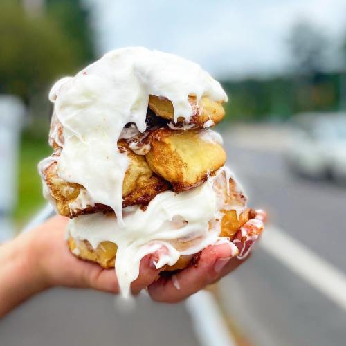 A hand holding a towering stack of cinnamon rolls drizzled with creamy icing against a blurred outdoor background.