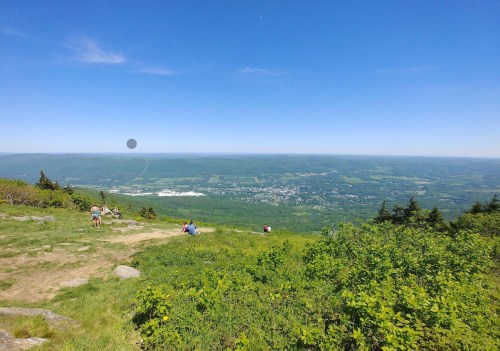A panoramic view from a mountain summit, showcasing green valleys and a clear blue sky with a few hikers in the foreground.