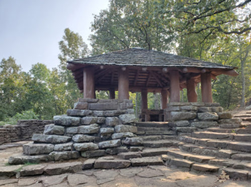 A rustic stone structure with a wooden roof, surrounded by trees and steps leading up to it.