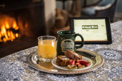 A cozy breakfast setting with a glass of juice, a coffee mug, and a plate of baked goods beside a tablet and a fireplace.