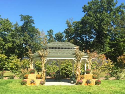 A white gazebo surrounded by colorful flowers, hay bales, and pumpkins under a clear blue sky.