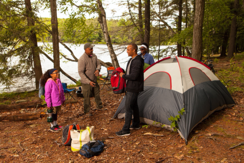A family sets up a tent by a lake, surrounded by trees, with camping gear and a cheerful atmosphere.