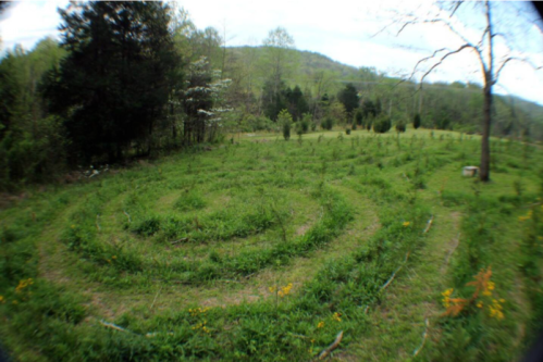 A grassy spiral maze surrounded by trees and hills under a cloudy sky.