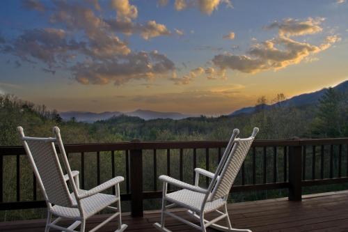 Two white rocking chairs on a wooden deck overlook a scenic mountain view at sunset, with clouds in the sky.