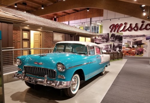A vintage turquoise and white car displayed in a museum setting with a modern backdrop and historical photos.
