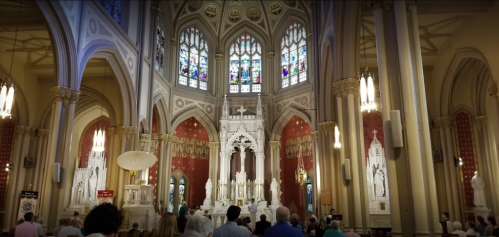 Interior of a grand church with stained glass windows, ornate altars, and a congregation gathered for a service.