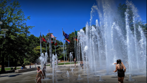 Children play in a splash pad with water fountains, surrounded by flags and trees under a clear blue sky.