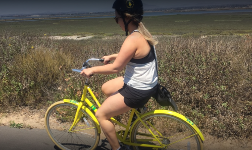 A person rides a yellow bicycle along a path with greenery and a body of water in the background.