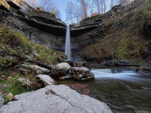 A serene waterfall cascades down rocky cliffs into a tranquil pool, surrounded by autumn foliage and trees.