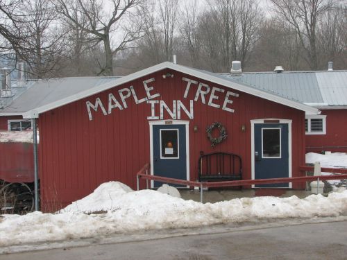 Red building with "Maple Tree Inn" sign, two doors, and a wreath, surrounded by snow and bare trees.
