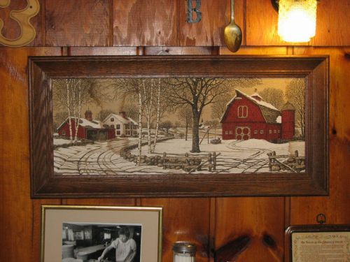 Framed artwork of a snowy farm scene with red barns, trees, and a wooden fence, displayed on a wooden wall.