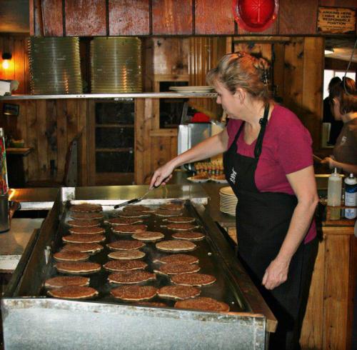 A woman in an apron cooks pancakes on a large griddle in a rustic kitchen setting.