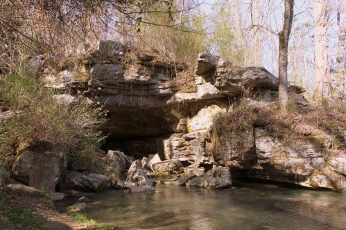 A rocky outcrop beside a calm stream, surrounded by trees and greenery in a serene natural setting.