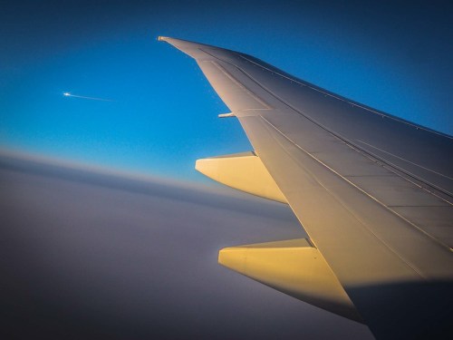 Airplane wing against a clear blue sky, with a distant jet contrail visible in the background.