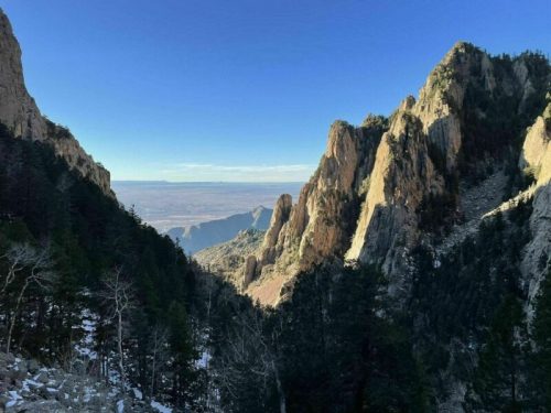 A scenic view of rugged mountains and a valley, framed by trees under a clear blue sky.
