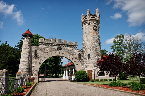 Stone archway with towers and clock, marking the entrance to Highland Lawn Cemetery, surrounded by trees and blue sky.
