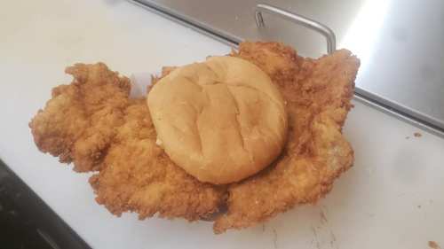 A large fried chicken fillet topped with a sandwich bun, served on a white surface.