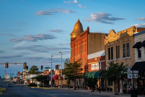 A quiet street scene featuring historic buildings, trees, and a blue sky with scattered clouds at sunset.
