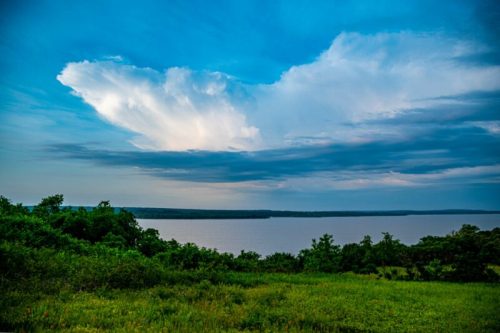 A serene landscape featuring a lake surrounded by greenery under a blue sky with fluffy clouds.