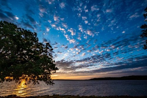 A serene lakeside scene at sunset, featuring vibrant clouds and a silhouette of a tree against the colorful sky.
