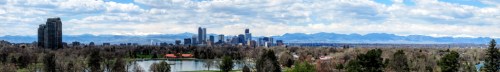 Panoramic view of a city skyline with mountains in the background, featuring a lake and green trees in the foreground.