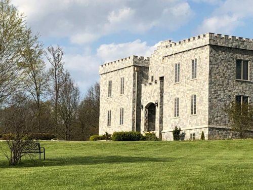 A stone castle with turrets sits on a grassy lawn, surrounded by trees under a partly cloudy sky.