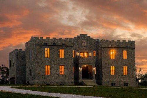 A stone castle with illuminated windows against a dramatic sunset sky.