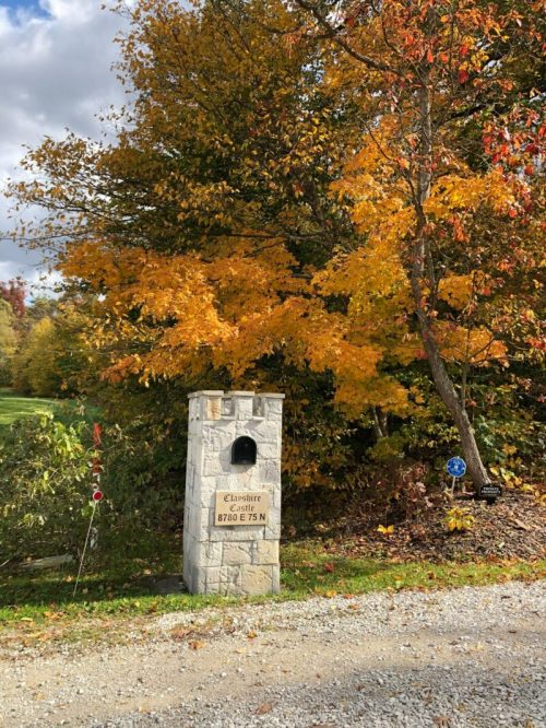 A stone mailbox stands beside a gravel road, surrounded by vibrant autumn foliage in shades of orange and yellow.