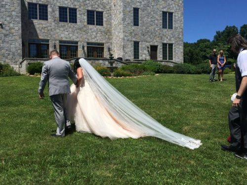A bride in a flowing gown and veil walks hand-in-hand with a groom on a sunny day outside a stone building.