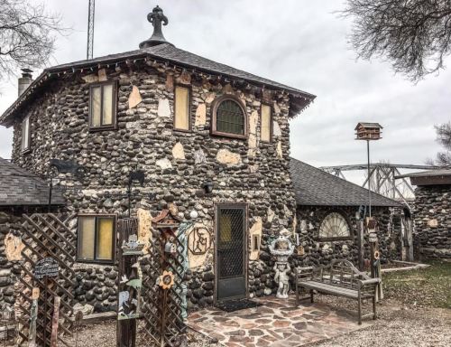 A unique stone house with a round tower, decorative elements, and a bench in front, set against a cloudy sky.