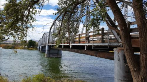 A metal bridge spans a river, surrounded by trees and a clear blue sky with scattered clouds.