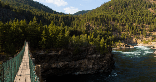 A wooden suspension bridge spans a river, surrounded by lush green forests and rocky cliffs under a blue sky.