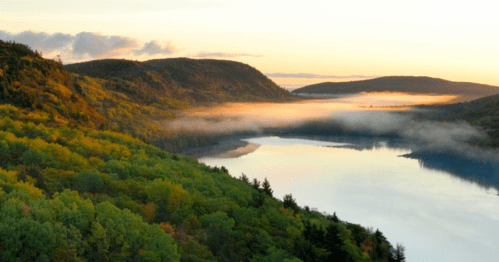 A serene landscape featuring a river surrounded by lush green hills and mist at sunrise.