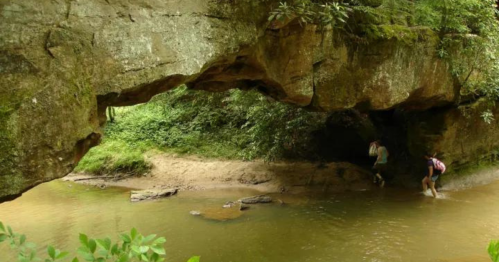 Two people explore a rocky area under a natural stone arch, with a calm stream and lush greenery surrounding them.