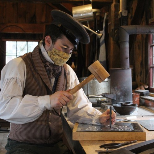 A craftsman in period attire uses a hammer and chisel to work on metal in a rustic workshop.