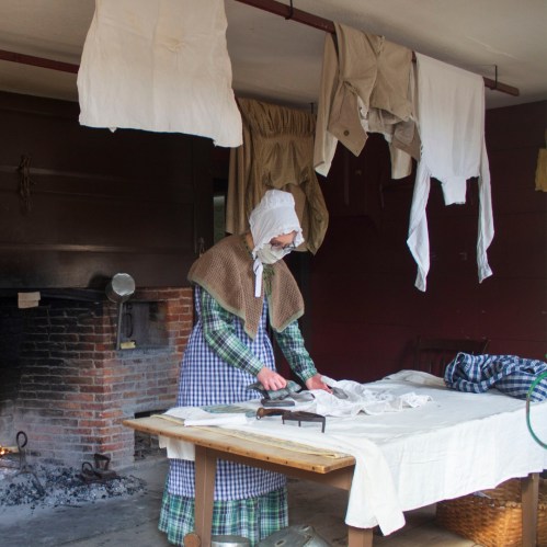 A woman in historical attire irons clothes on a table, with laundry hanging above in a rustic setting.