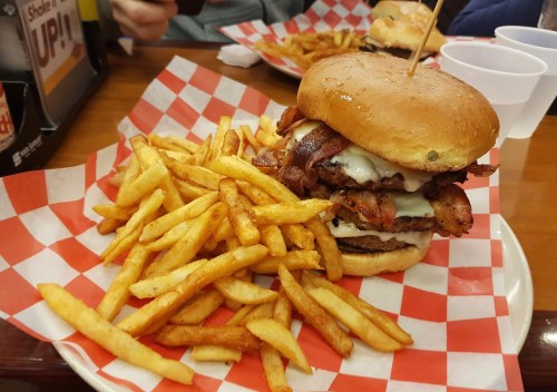 A large burger stacked with bacon and cheese, served with a side of crispy French fries on a checkered tablecloth.