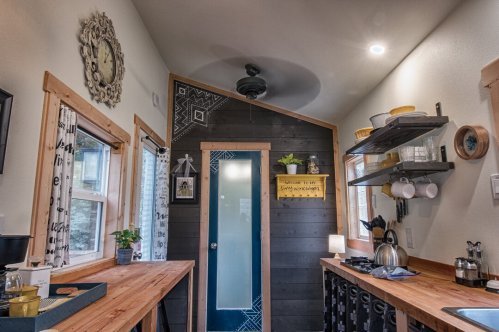 Cozy kitchen with wooden tables, shelves, and modern decor, featuring a blue door and natural light from windows.
