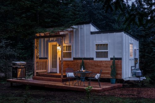 A cozy cabin with a wooden deck, grill, and outdoor seating, surrounded by trees in a serene setting at dusk.