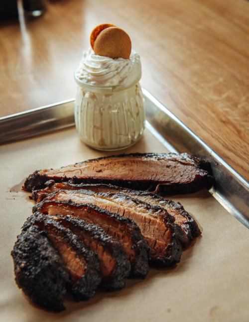 Sliced smoked brisket on parchment paper with a jar of whipped cream topped with a cookie in the background.