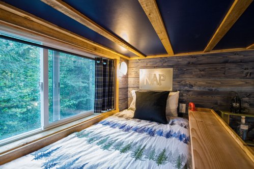 Cozy bedroom with a bed, blue ceiling, wooden walls, and a window overlooking trees, featuring a "NAP" sign.