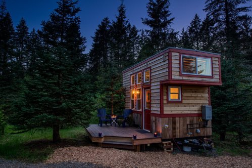 A cozy tiny house with large windows, surrounded by tall trees, on a wooden deck with outdoor seating at dusk.