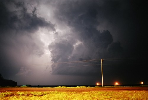 Dramatic dark clouds illuminated by lightning, with a power line and distant lights in a rural landscape at night.