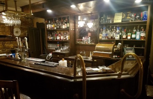 A cozy bar with wooden decor, shelves lined with various liquor bottles, and a vintage cash register.