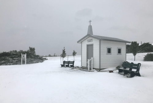 A small white chapel with a cross, surrounded by snow, with benches nearby and a cloudy sky above.