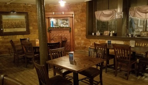 Cozy restaurant interior with wooden tables and chairs, stone walls, and a bar in the background. Warm lighting.