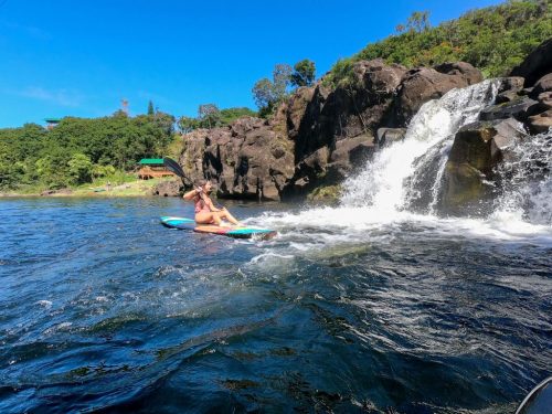 A person kayaking near a rocky waterfall on a sunny day, surrounded by lush greenery.