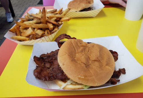 A close-up of a bacon sandwich on a yellow table, with a side of crispy fries in the background.