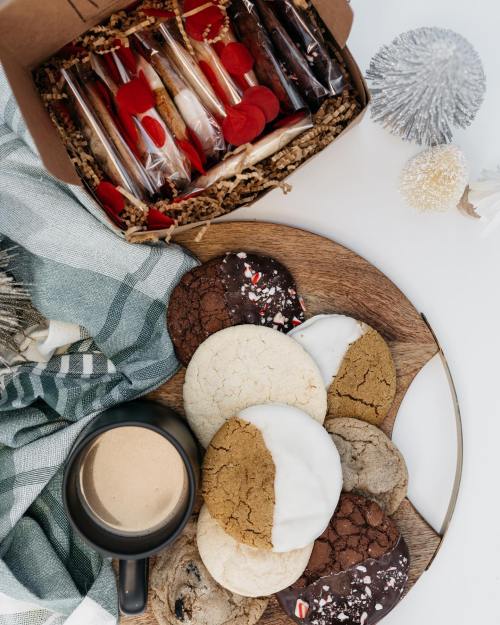 A wooden platter with assorted cookies, a coffee cup, and a gift box filled with treats, set on a cozy blanket.
